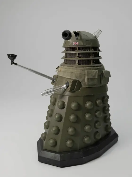 Full-size khaki Dalek replica with a Union Jack on the dome, canvas webbing and pouches around the shoulders, and its plunger arm and gun extended